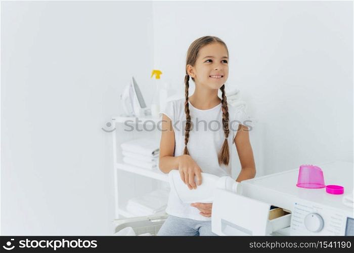 Little happy girl has two pigtails fills in washing machine with liquid detergent, pours into tray of washer, stands in laundry room with white walls, helps mother with washing, wears white t shirt