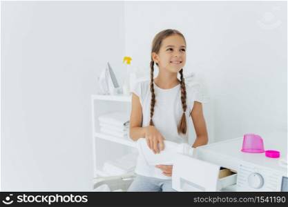 Little happy girl has two pigtails fills in washing machine with liquid detergent, pours into tray of washer, stands in laundry room with white walls, helps mother with washing, wears white t shirt