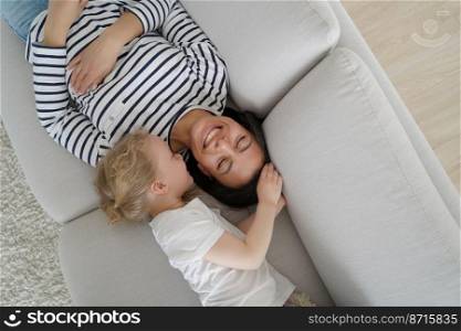 Little kid daughter hugging mother, lying on couch at home. Top view of smiling young mom and small preschooler girl enjoying touching moment together, playing, having fun on cozy sofa.. Little kid girl daughter hugging mother, lying on couch at home. Touching moment of motherhood