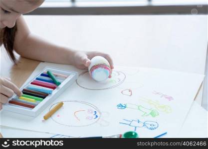 Little kid girl choosing crayon color to decorate Easter egg in hand. Cute child prepare for Easter holiday at home. Children education by drawing and painting