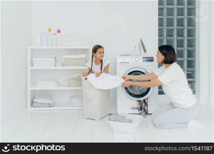 Little kid helps mum with washing, sits in basket with laundry. Busy housewife does housework, washes laundry in washing machine, loads washer, pose in spacious white bathroom. Household concept