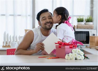 Little mixed race daughter kissing young Asian dad on cheek while giving and opening gift box and card together. Family sitting at home kitchen celebrating father day or birthday special occasion
