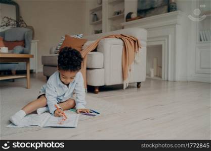 Little mixed race kid boy drawing in coloring book with lots of different felt tip pen while sitting alone on floor in middle of living room at home. Children leisure and developing activities. Little mixed race kid boy drawing in coloring book while spending leisure time alone at home