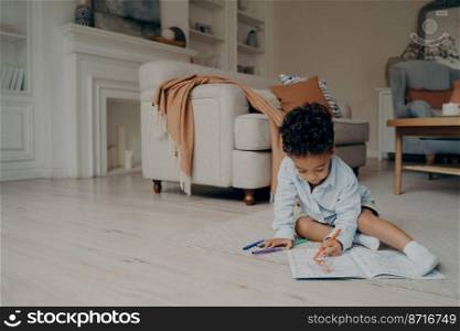 Little mixed race kid boy drawing in coloring book with lots of different felt tip pen while sitting alone on floor in middle of living room at home. Children leisure and developing activities. Little mixed race kid boy drawing in coloring book while spending leisure time alone at home