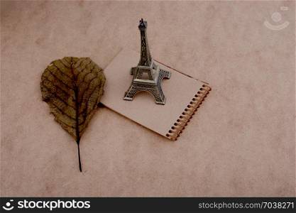 Little model Eiffel Tower, notebook and a dry leaf on brown background