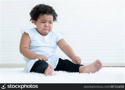 Little pitiful African American chubby kid girl is crying with tears drop from eyes while sitting on fluffy carpet on floor at home. Child emotion care concept. White background. Copy space