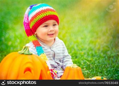 Little pretty child sitting on green grass field near two cute pumpkins with carved faces, playing with festive decoration, celebrating Halloween holiday