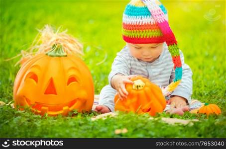 Little pretty child sitting on the green grass field near two cute pumpkins with carved faces, celebrating Halloween holiday