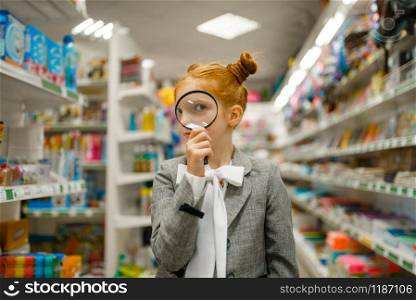 Little school girl looks through magnifying glass, shopping in stationery store. Little school girl looks through magnifying glass
