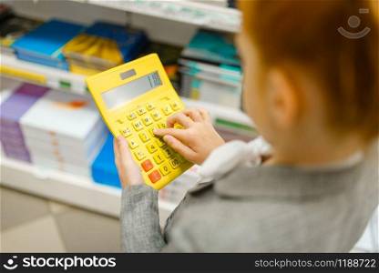 Little schoolgirl holds yellow calculator, shopping in stationery store. Female child buying office supplies in shop, schoolchild in supermarket. Schoolgirl holds calculator, stationery store