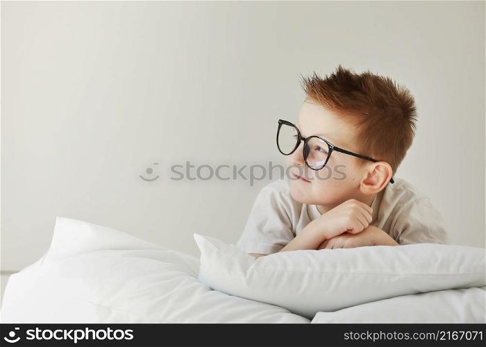 Little smiling boy in glasses is lying on white bed with pillows. Copy Space. Little smiling boy in glasses is lying on white bed with pillows. Copy Space.