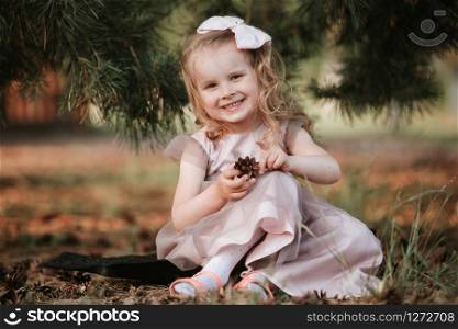 little smiling girl with a bow on her head and in a dress with sits under a tree. Child enjoys playing with cones.. little smiling girl with a bow on her head and in a dress with sits under a tree. Child enjoys playing with cones