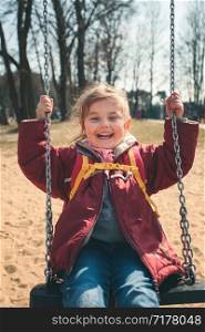 Little smilling happy girl swinging in a park on sunny spring day. Child looking at camera wearing red jacket