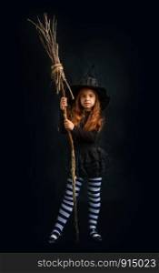 little witch girl in traditional Halloween dress and pointed hat is standing with a wooden whisk on a dark background. little girl witch