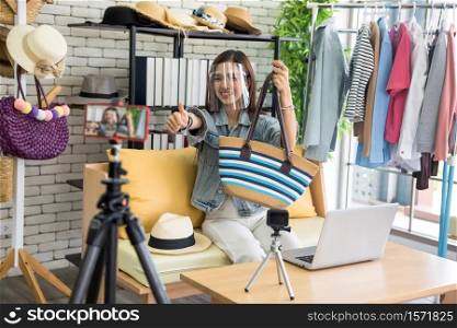 Live streaming content for selling fashion bag online by smartphone on tripod. Beauty blogger or internet influencer. Beautiful young Asian woman vlogger blogger with face shield show handbag on social media.