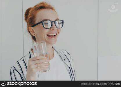 Living healthy lifestyle. Happy laughing ginger woman standing in kitchen in morning with glass of pure mineral water, feeling thirsty dehydrated while spending time at home, taking care of herself. Happy laughing ginger woman standing in kitchen in morning with glass of pure mineral water
