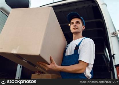 Loader in uniform holding carton box, delivery service. Man standing at cardboard packages in vehicle, male deliver, courier or shipping job