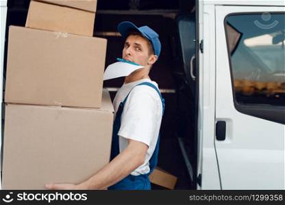 Loader in uniform holding stack of parcels, delivery service. Man standing at cardboard packages in vehicle, male deliver, courier or shipping job