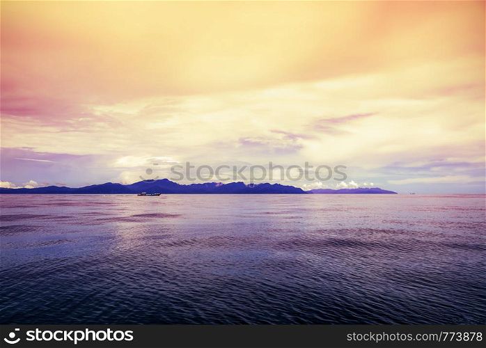 Local passenger boat are sailing on the water surface in the sea with a silhouette of the island in the background. under the bright red yellow sky during the sunset at Pak Bara bay, Satun, Thailand. Sea under the bright sky during the sunset at Pak Bara bay, Thailand