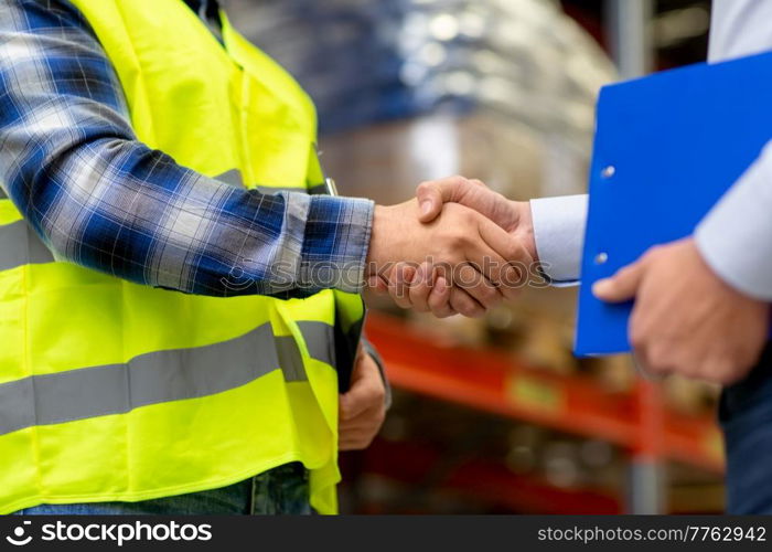 logistic business and cooperation concept - close up of manual worker and businessman with clipboard shaking hands and making deal at warehouse. worker and businessman shaking hands at warehouse