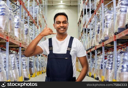 logistic business and people concept - happy smiling indian loader or worker making phone call gesture over warehouse background. worker making phone call gesture at warehouse