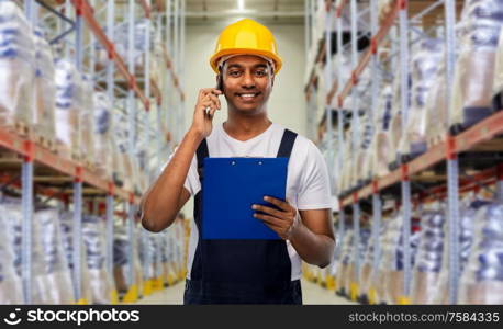 logistic business and people concept - happy smiling indian loader or worker in helmet with clipboard calling on smartphone over warehouse background. indian worker calling on smartphone at warehouse