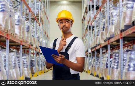 logistic business and people concept - happy smiling indian loader or worker in helmet with clipboard and pencil over warehouse background. thinking indian worker with clipboard at warehouse