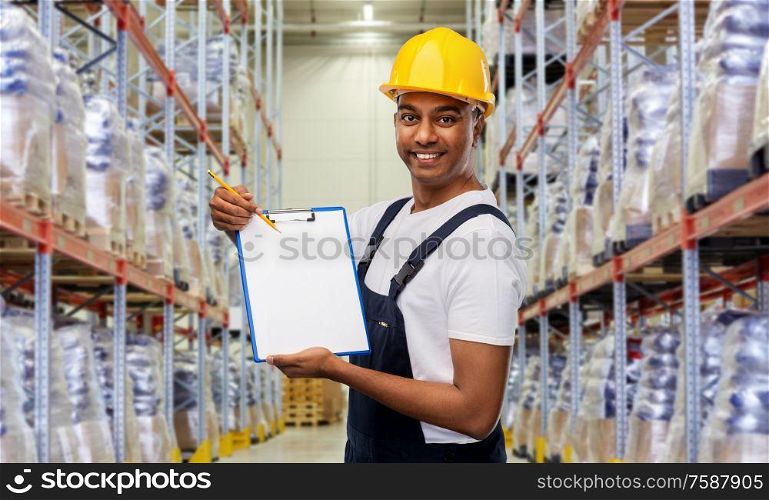 logistic business and people concept - happy smiling indian loader or worker in helmet with clipboard and pencil over warehouse background. happy indian worker with clipboard at warehouse
