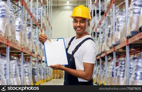 logistic business and people concept - happy smiling indian loader or worker in helmet with clipboard and pencil over warehouse background. happy indian worker with clipboard at warehouse