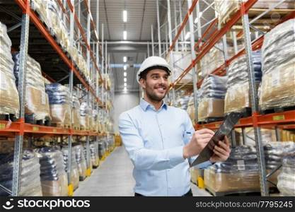 logistic business, shipment and people concept - businessman in helmet with clipboard checking goods at warehouse. businessman in helmet with clipboard at warehouse