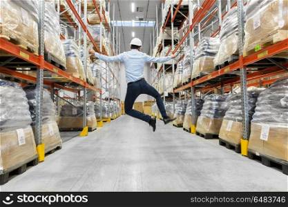 logistic business, success and people concept - happy businessman in helmet jumping at warehouse. happy businessman in helmet jumping at warehouse. happy businessman in helmet jumping at warehouse