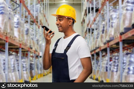 logistic business, technology and people concept - happy smiling indian loader or worker in helmet using voice command recorder on smartphone over warehouse background. happy indian loader recording voice on smartphone