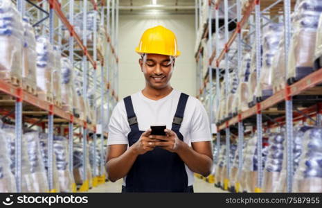 logistic business, technology and people concept - happy smiling indian loader or worker in helmet with smartphone over warehouse background. loader or worker with smartphone at warehouse