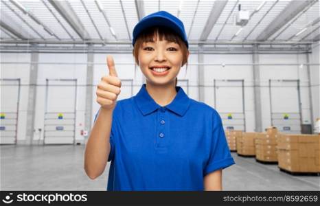 logistics, shipping and job concept - happy smiling delivery woman in blue uniform showing thumbs up gesture over warehouse background. delivery woman showing thumbs up at warehouse