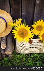 Logs, a hat, a bouquet of sunflowers in a straw bag are standing near a wooden house. Close-up.. Logs, a hat, a bouquet of sunflowers in a straw bag are standing near a wooden house.