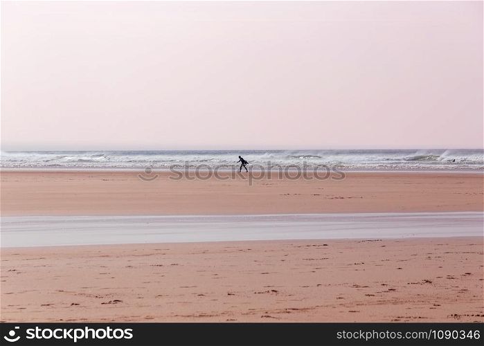 Lone surfer, carrying a surfboard, walking along the beach in Cornwall, UK