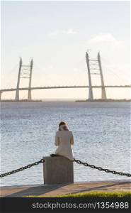 Loneliness, desire to be alone, walk along the water. Woman sitting by sea, looking at the bridge. View from the back.