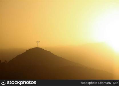 Lonely cross on hill yellow orange, surrounded by setting sun