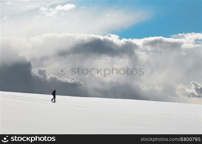 Lonely figure of man walking down a hill. White clear surface of snow till flat horizon and beautiful cloudscape, gloomy sky above a man; copy space. Concept of hiking and tourism.
