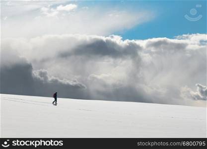 Lonely figure of man walking down a hill. White clear surface of snow till flat horizon and beautiful cloudscape, gloomy sky above a man; copy space. Concept of hiking and tourism.
