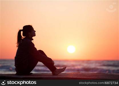 Lonely girl sitting on the sunrise beach. Woman silhouette over sunrise sky