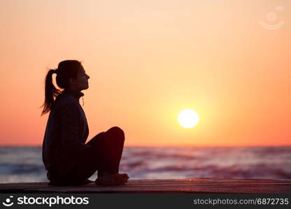 Lonely girl sitting on the sunrise beach. Woman silhouette over sunrise sky