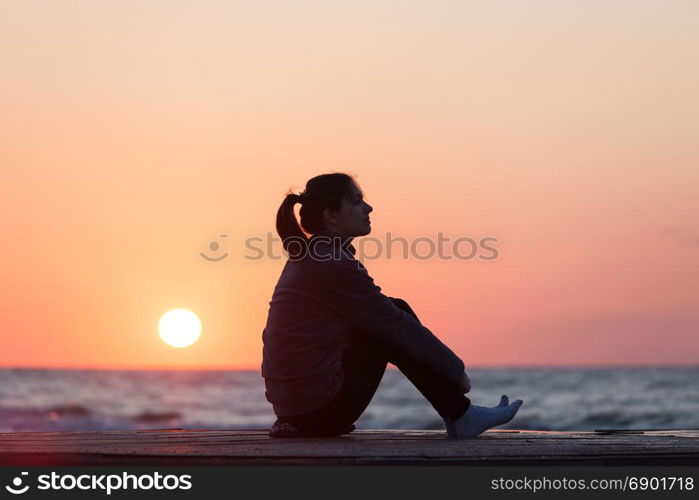 Lonely girl sitting on the sunrise beach. Woman silhouette over sunrise sky