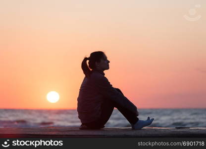 Lonely girl sitting on the sunrise beach. Woman silhouette over sunrise sky