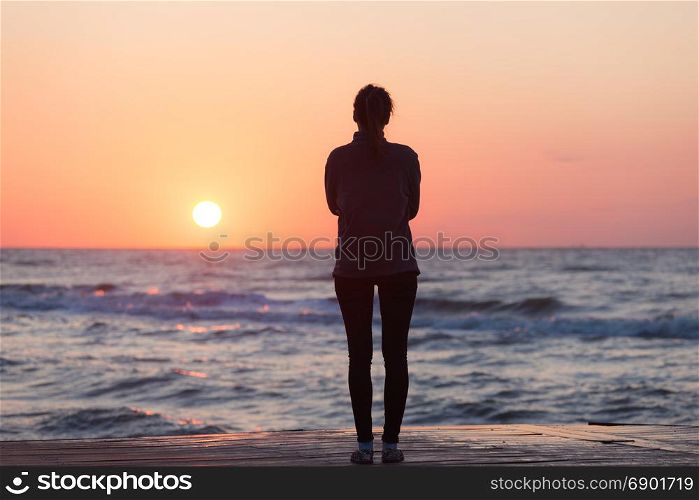 Lonely girl standing on the sunrise beach. Woman silhouette over sunrise sky