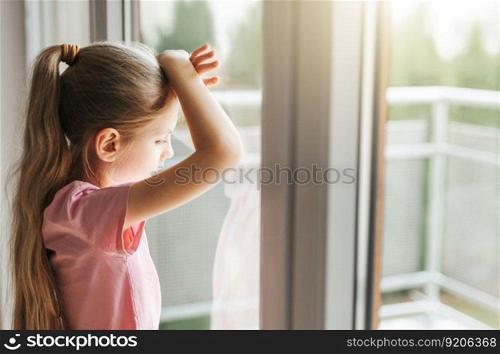 Lonely little girl  looking out window, standing at home alone. 