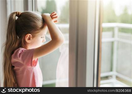 Lonely little girl  looking out window, standing at home alone. 