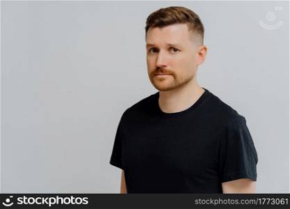 Lonely mood. Studio shot of unhappy sad ginger bearded man looking at camera with worried face expression, feeling upset while standing in black shirt against grey wall. People and unpleasant emotions. Young ginger man feeling sad and disappointed, looking at camera with frustrated face expression