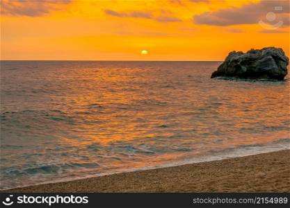 Lonely rock in the sea near the sandy beach. Colorful sunrise over the calm waters of the Greek Mediterranean coast. Lonely Rock in the Sea at Dawn and Strip of Sandy Beach