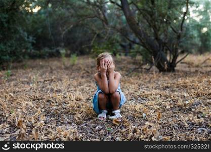 Lonely sad little girl squatting in the forest, covers her face with her hands Feelings concept.